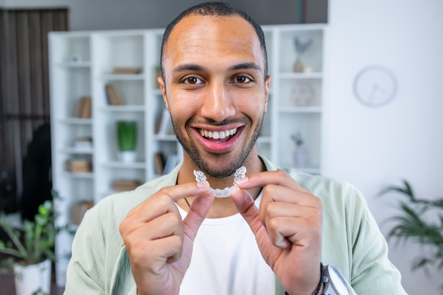 Man smiling and holding a clear aligner – Invisalign San Mateo Happy man showing a clear Invisalign aligner indoors – Invisalign San Mateo