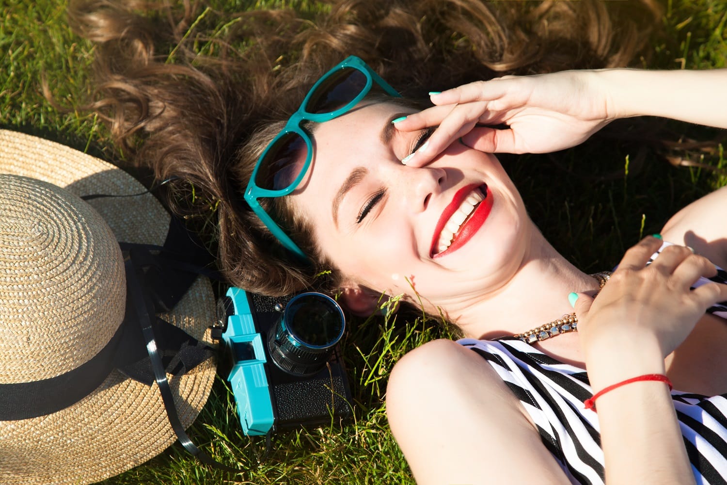 Smiling Woman Relaxing on Grass with Sunglasses and Hat – Cosmetic Dentist San Mateo Happy woman lying on the grass with bright red lipstick, turquoise sunglasses, and a sun hat beside her, showing off a radiant smile. – Cosmetic Dentist San Mateo