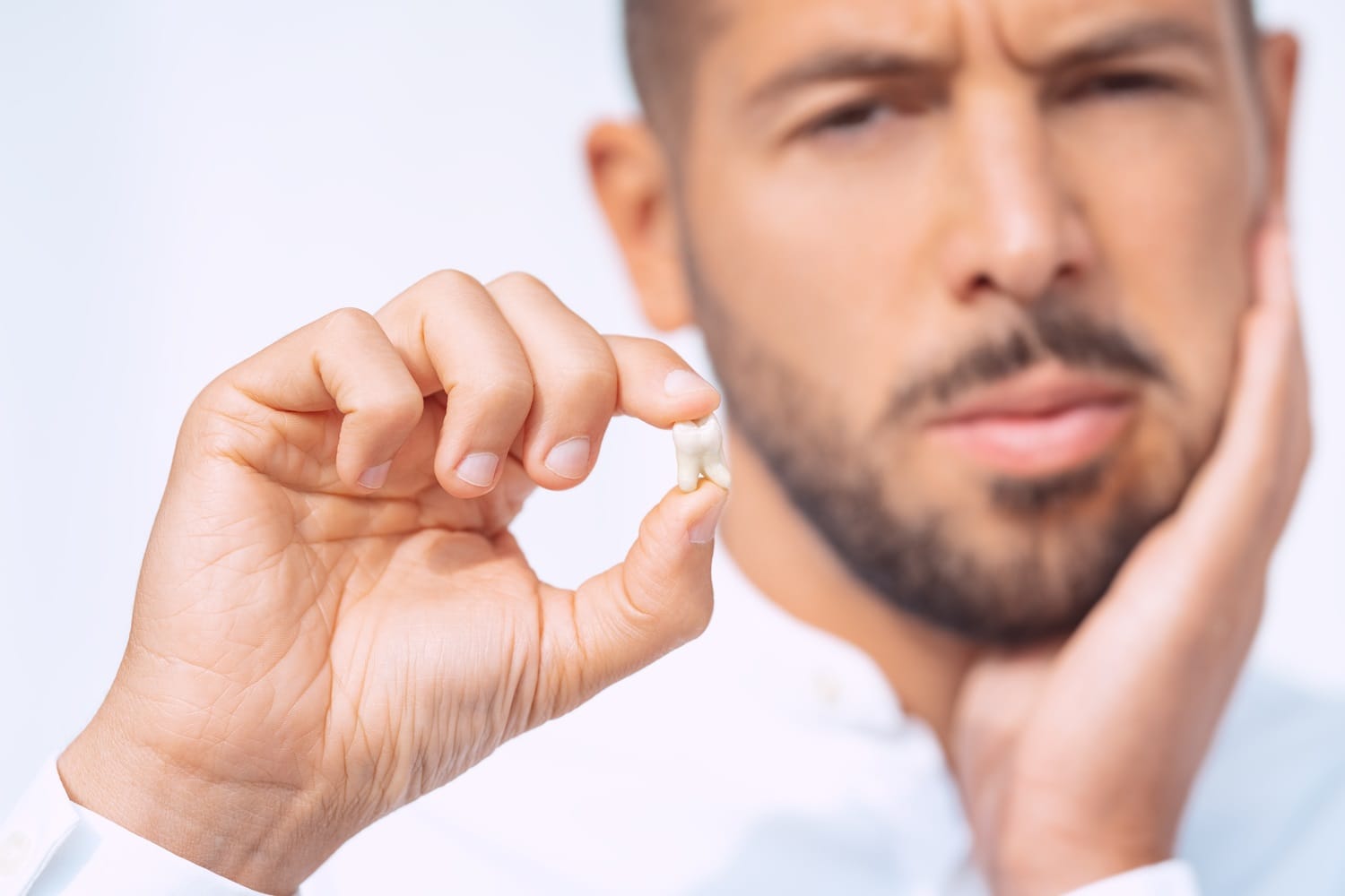 A close-up of a man holding an extracted wisdom tooth between his fingers while pressing his other hand to his cheek in discomfort – Wisdom Teeth Removal San Mateo.