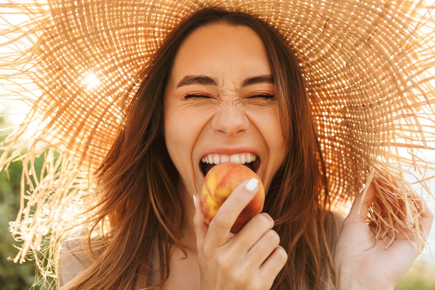 Woman Smiling and Biting a Peach Under Straw Hat – Cosmetic Dentist San Mateo Joyful woman wearing a sun hat and smiling brightly while biting into a peach, representing a confident and healthy smile. – Cosmetic Dentist San Mateo