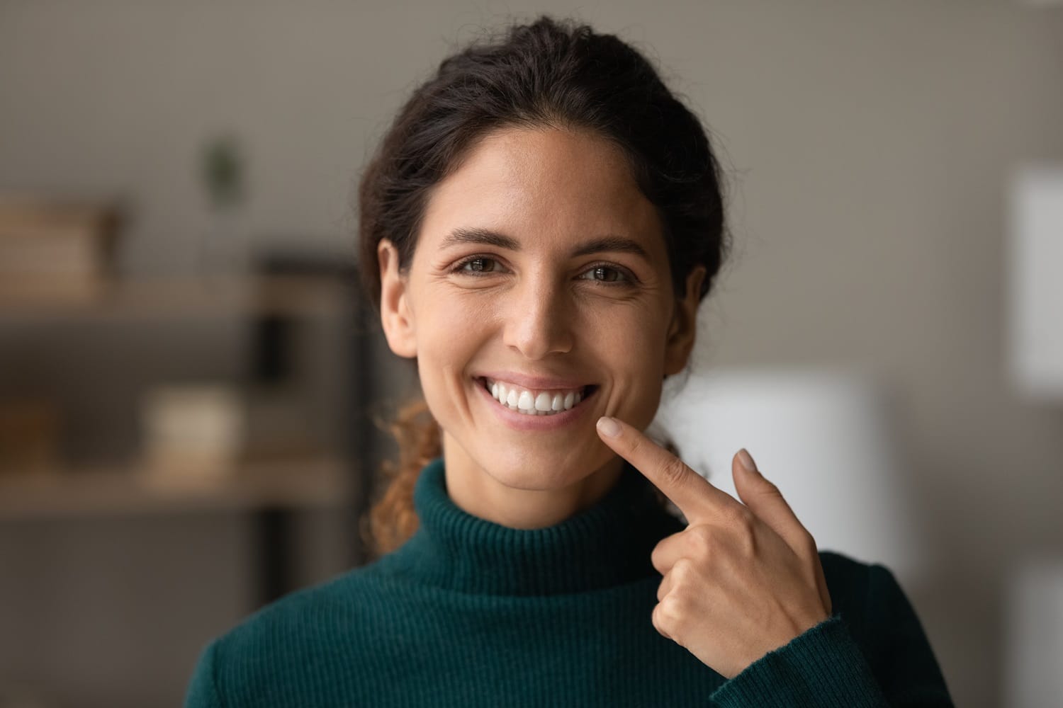 Confident Woman Pointing at Her Bright Smile – Cosmetic Dentist San Mateo Smiling woman wearing a green sweater and pointing at her white teeth, showcasing the results of professional cosmetic dental care. – Cosmetic Dentist San Mateo