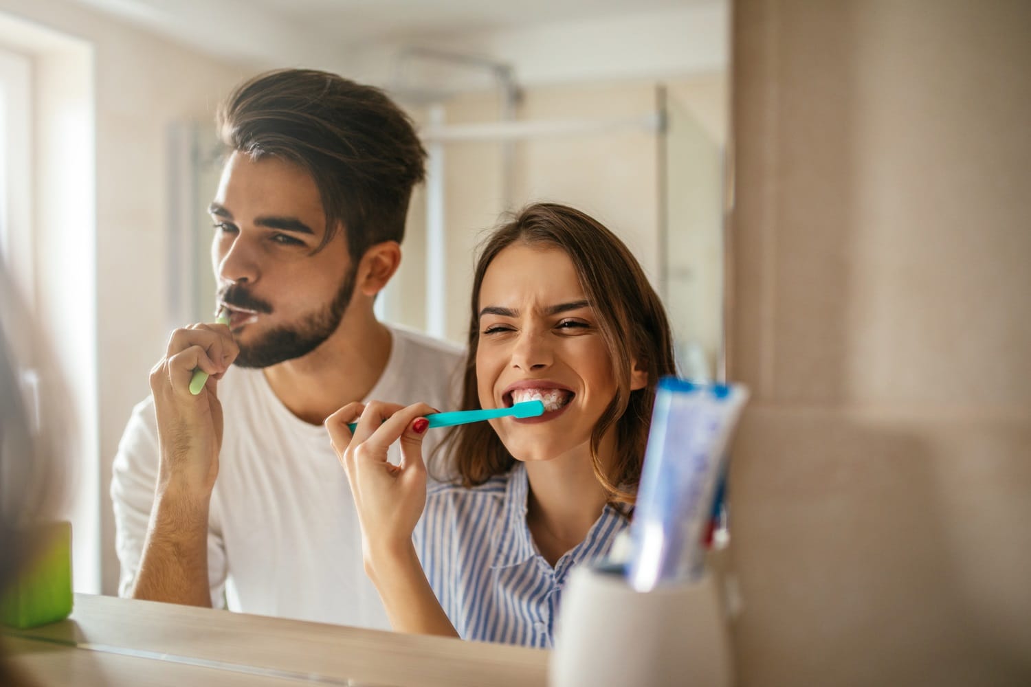 Couple Brushing Teeth Together — Dentist in San Mateo Smiling couple brushing their teeth in front of a bathroom mirror, promoting good oral hygiene habits encouraged by dentists in San Mateo
