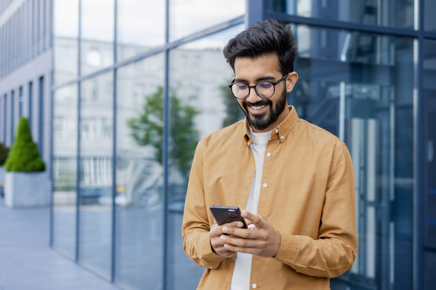 Smiling Man Using Smartphone — Dentist in San Mateo A man in glasses and a mustard-colored shirt smiles while using his smartphone outside a modern glass building - Dentist in San Mateo