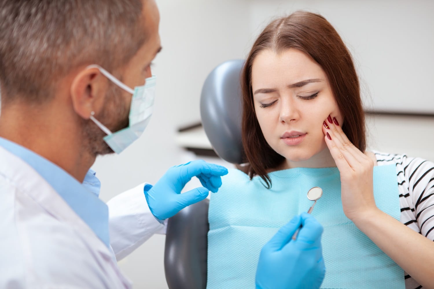 Woman Experiencing Tooth Pain at Dentist – Emergency Dentist in San Mateo Female patient sitting in a dental chair holding her cheek in pain while a dentist examines her, representing urgent dental care. – Emergency Dentist in San Mateo