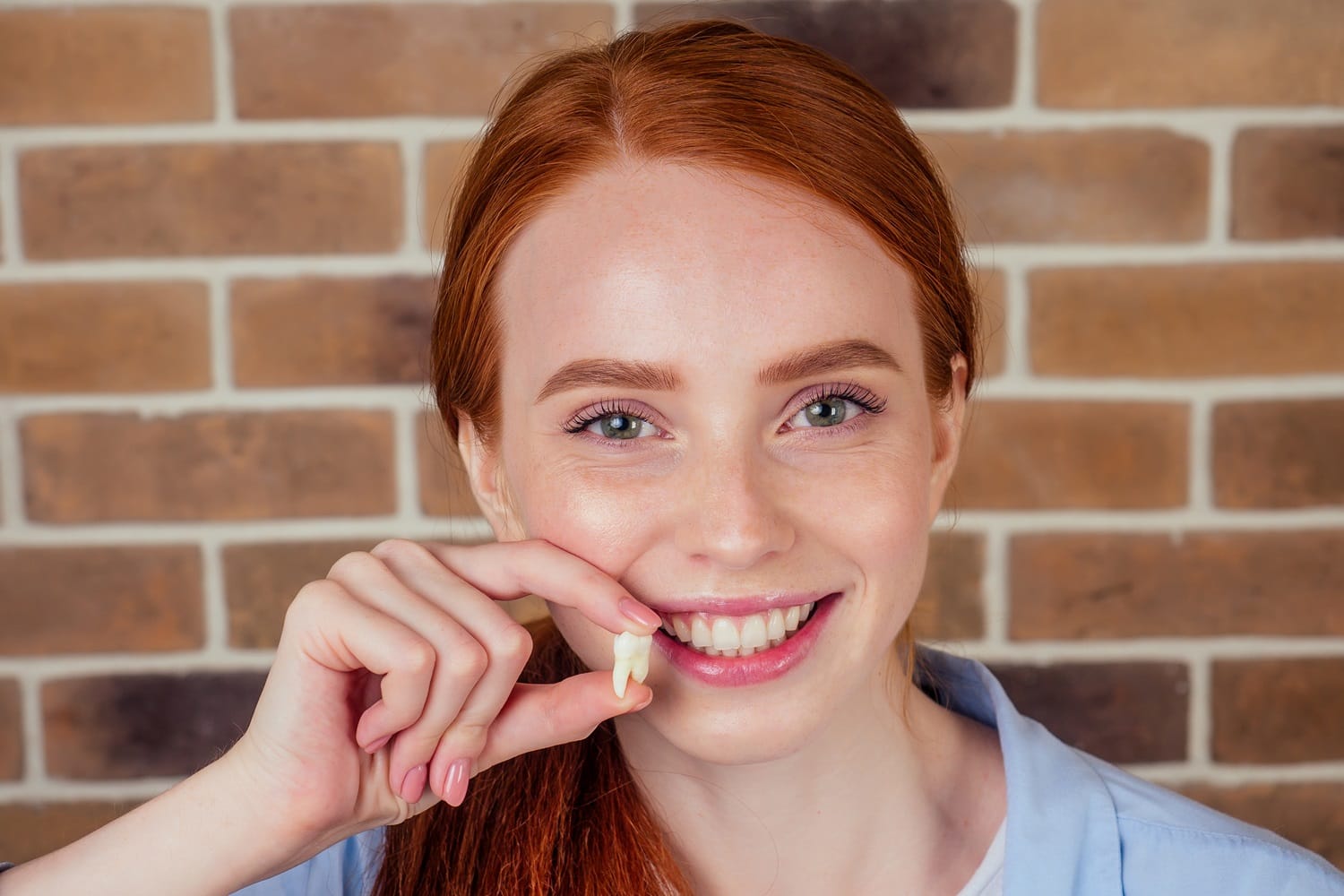 A cheerful young woman with red hair smiling and holding up a removed wisdom tooth in front of a brick wall – Wisdom Teeth Removal San Mateo