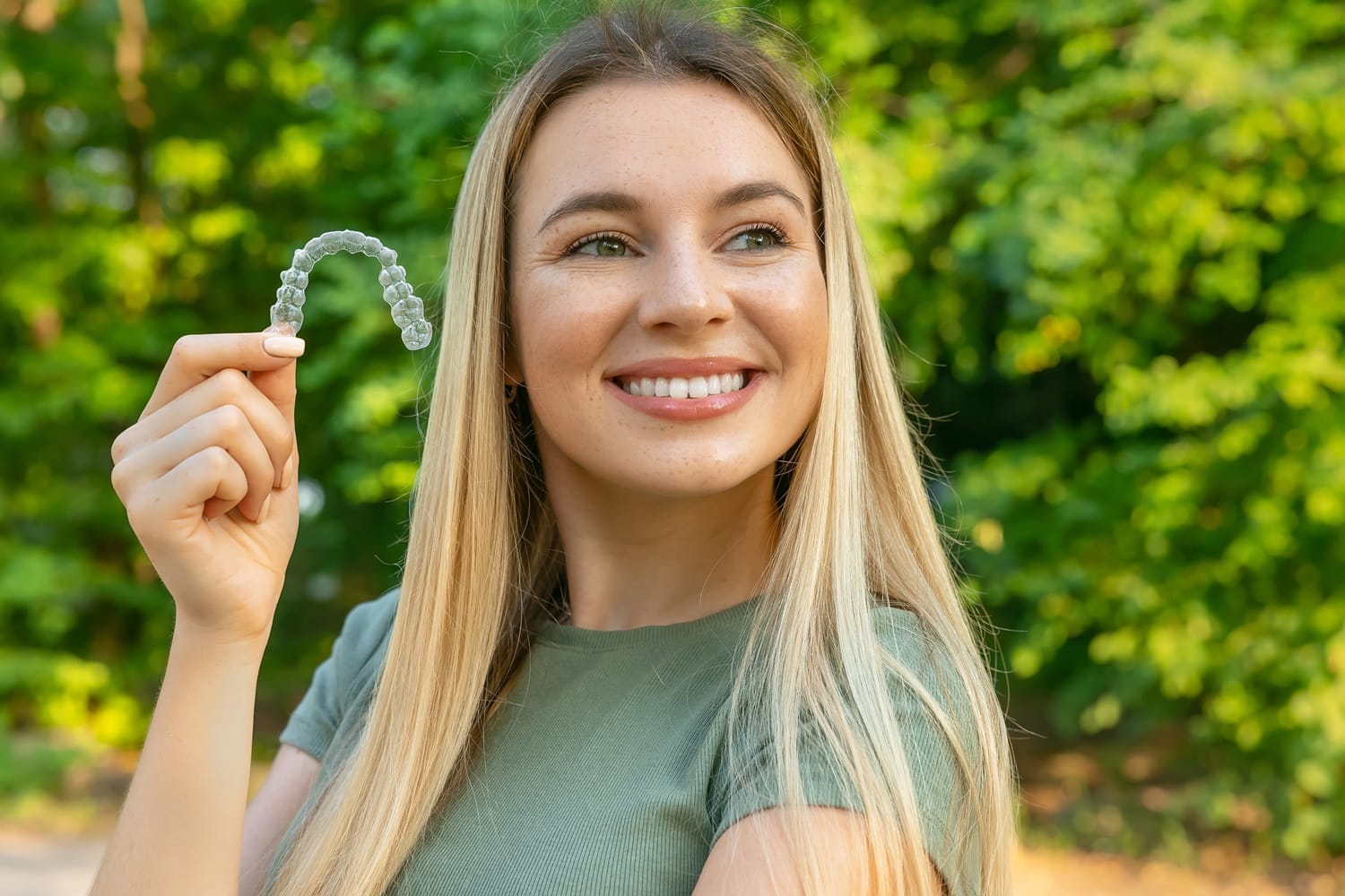 Woman smiling and holding a clear aligner – Invisalign San Mateo Happy woman outdoors showing a clear Invisalign aligner – Invisalign San Mateo